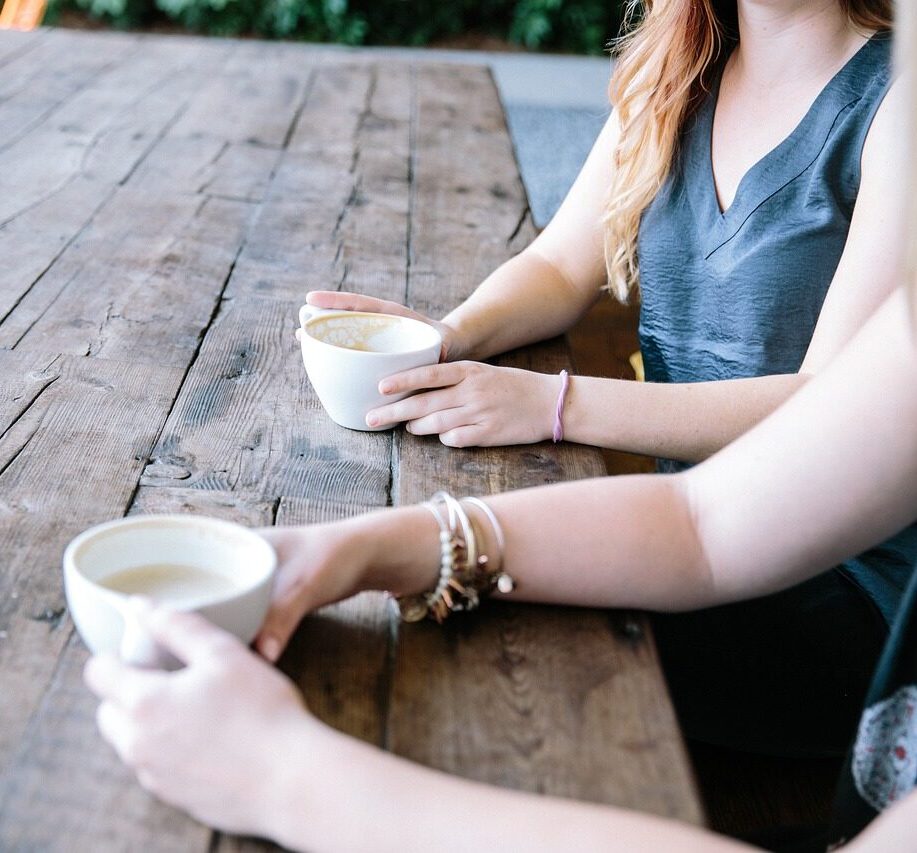 Zoomed in picture of 2 people sitting at a wooden outdoor table drinking coffee chatting like you might in a coaching session. CHCNZA coaches are warm friendly people.