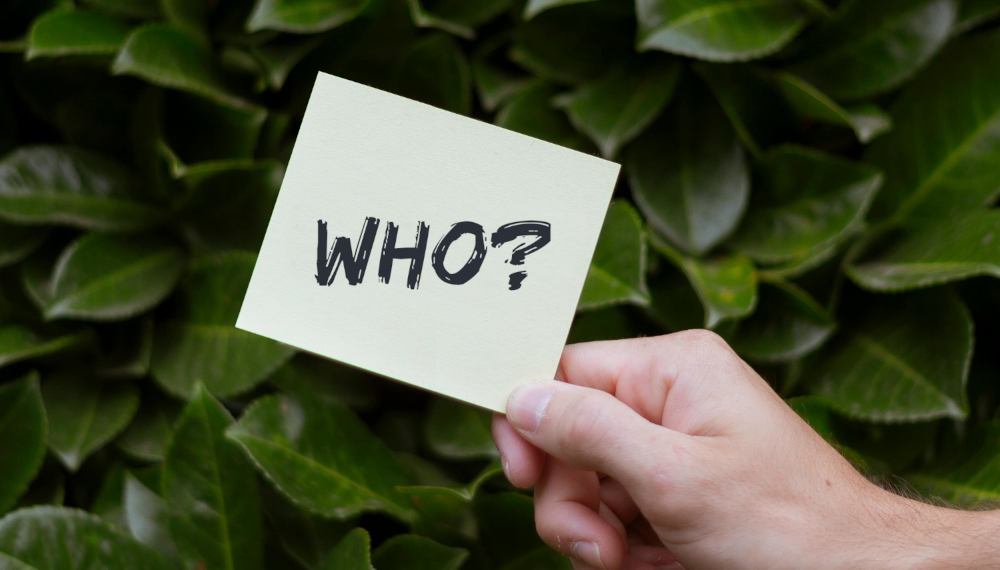 Person holding post it card in front of leaves with the word who?