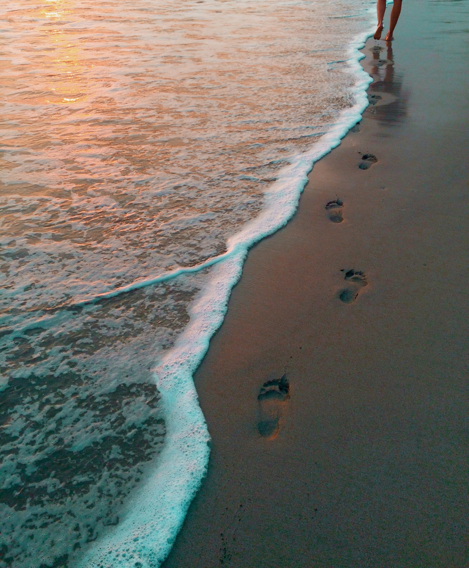 a person walking along a sandy beach with wave washing over the footprints.