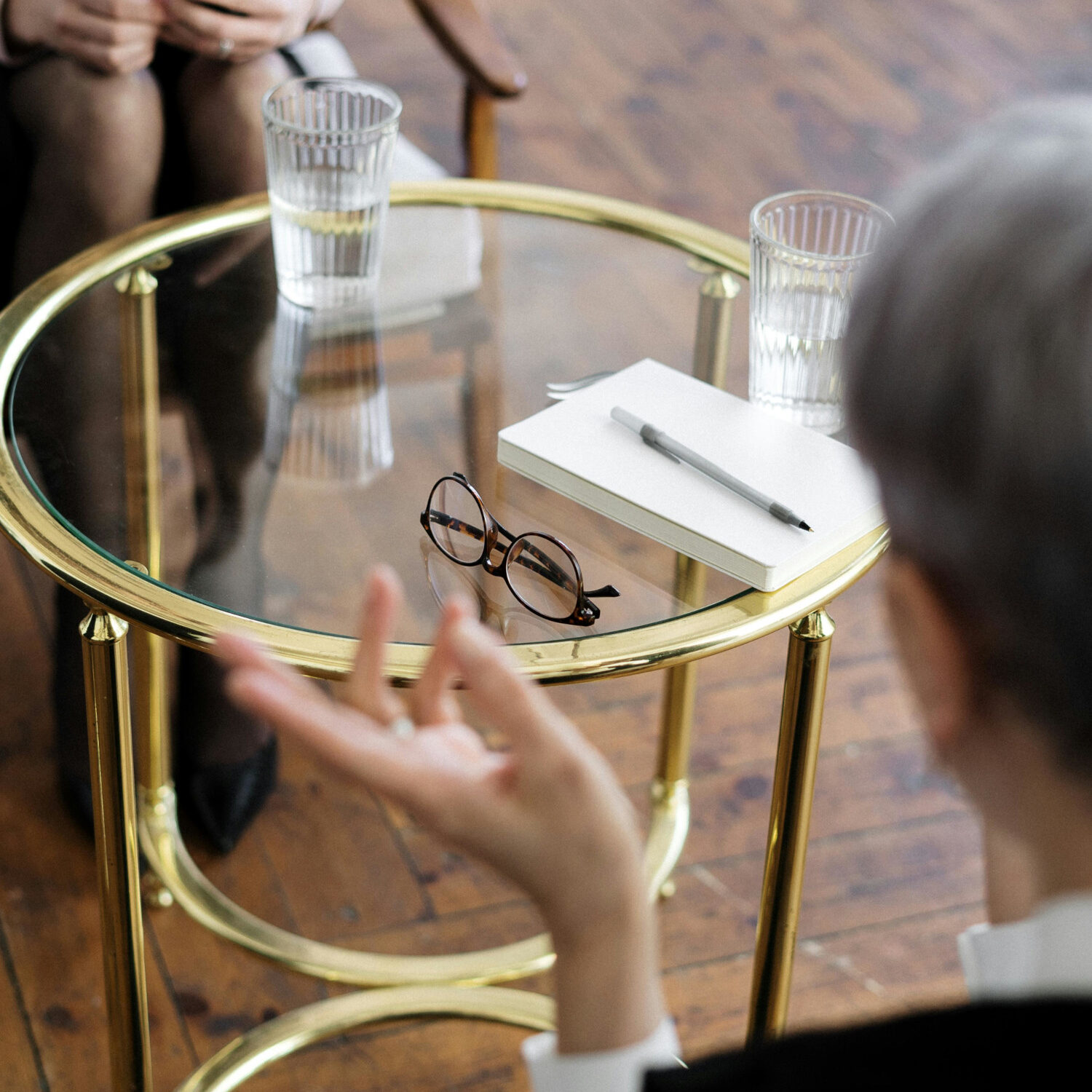 A coach coaching a client. Sitting opposite each other. Table in the middle with 2 glasses of water, notepad and pen, and a pair of glasses. Showing what a coaching session could look like